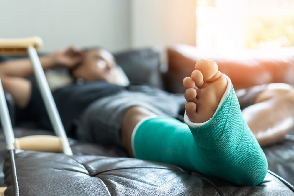 Man resting on couch with leg in green cast and crutches nearby, sunlight through window.