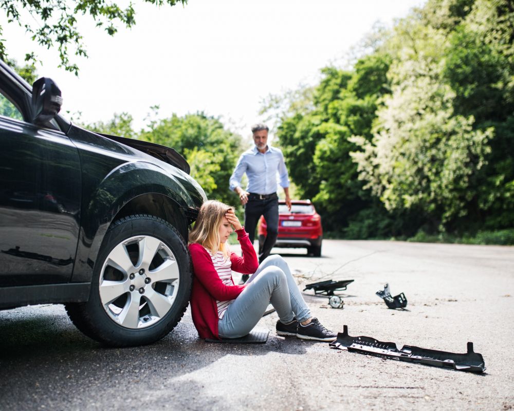 Injured woman sitting beside a damaged car after a hit-and-run accident while another driver approaches.
