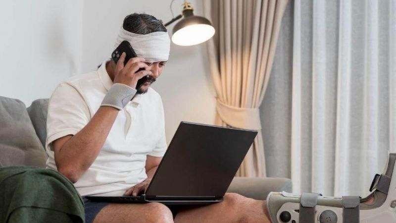 Man with a bandaged head and a leg in a cast sits on a sofa, talking on a phone while using a laptop nearby at home.