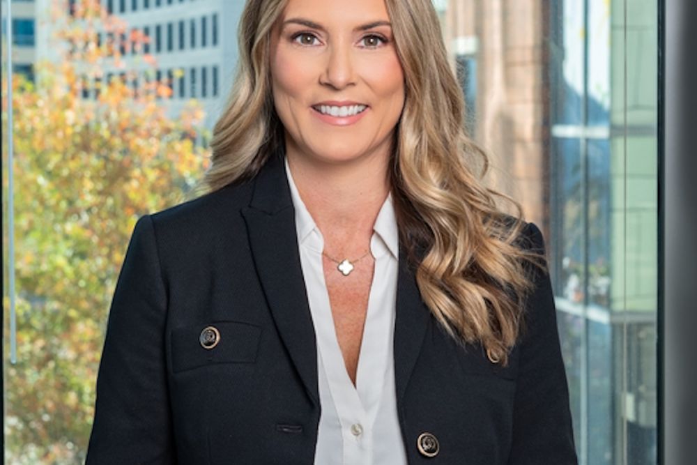 Professional woman smiling in formal attire with cityscape background, standing near a window in a modern office.