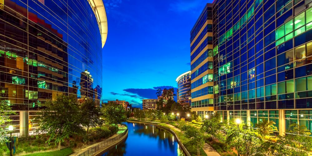 Modern glass buildings reflecting in a canal with trees and pathway at dusk.