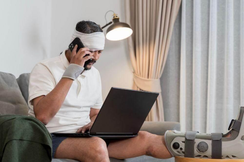 Man with a bandaged head and a leg in a cast sits on a sofa, talking on a phone while using a laptop nearby at home.