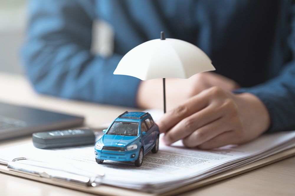 Blue toy car under umbrella on insurance documents, symbolizing vehicle protection and coverage, beside a laptop.