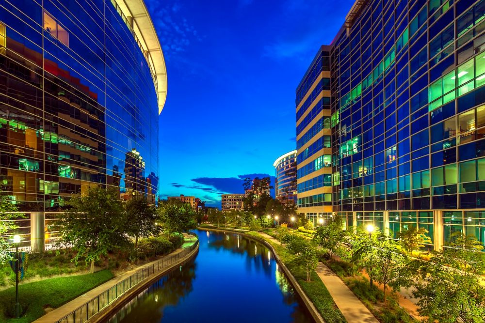 Modern glass buildings reflecting in a canal with trees and pathway at dusk.