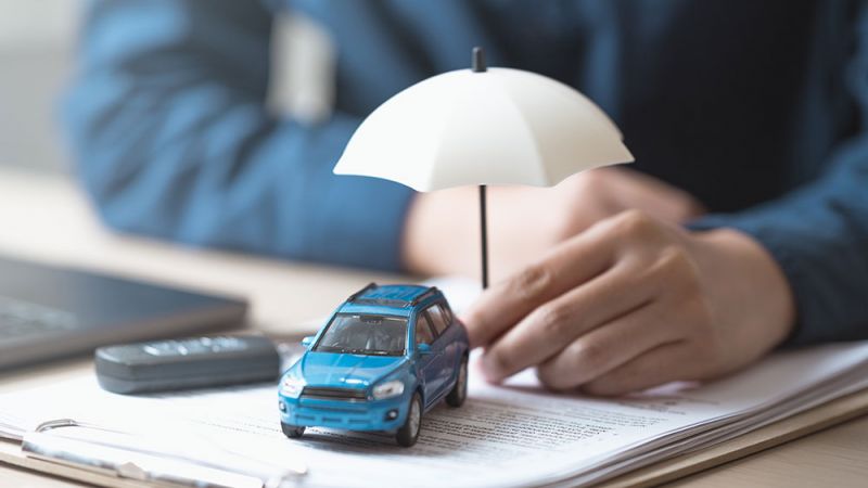 Blue toy car under umbrella on insurance documents, symbolizing vehicle protection and coverage, beside a laptop.