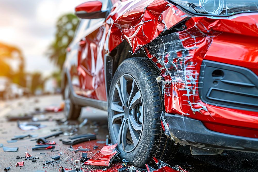 Red car with severe side damage after collision, shattered glass and debris on road in bright daylight.
