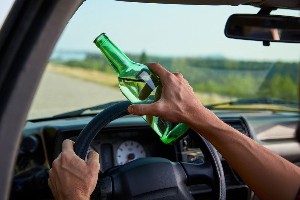 Person holding green bottle while driving, emphasizing dangers of drinking and driving on the road.