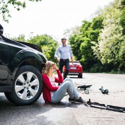Injured woman sitting beside a damaged car after a hit-and-run accident while another driver approaches.