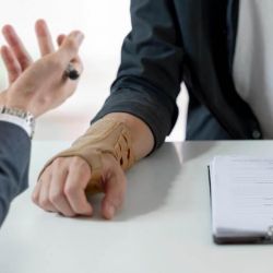 Handshake across a table between two people, one with a forearm in a beige cast, a pen and forms on a clipboard nearby.