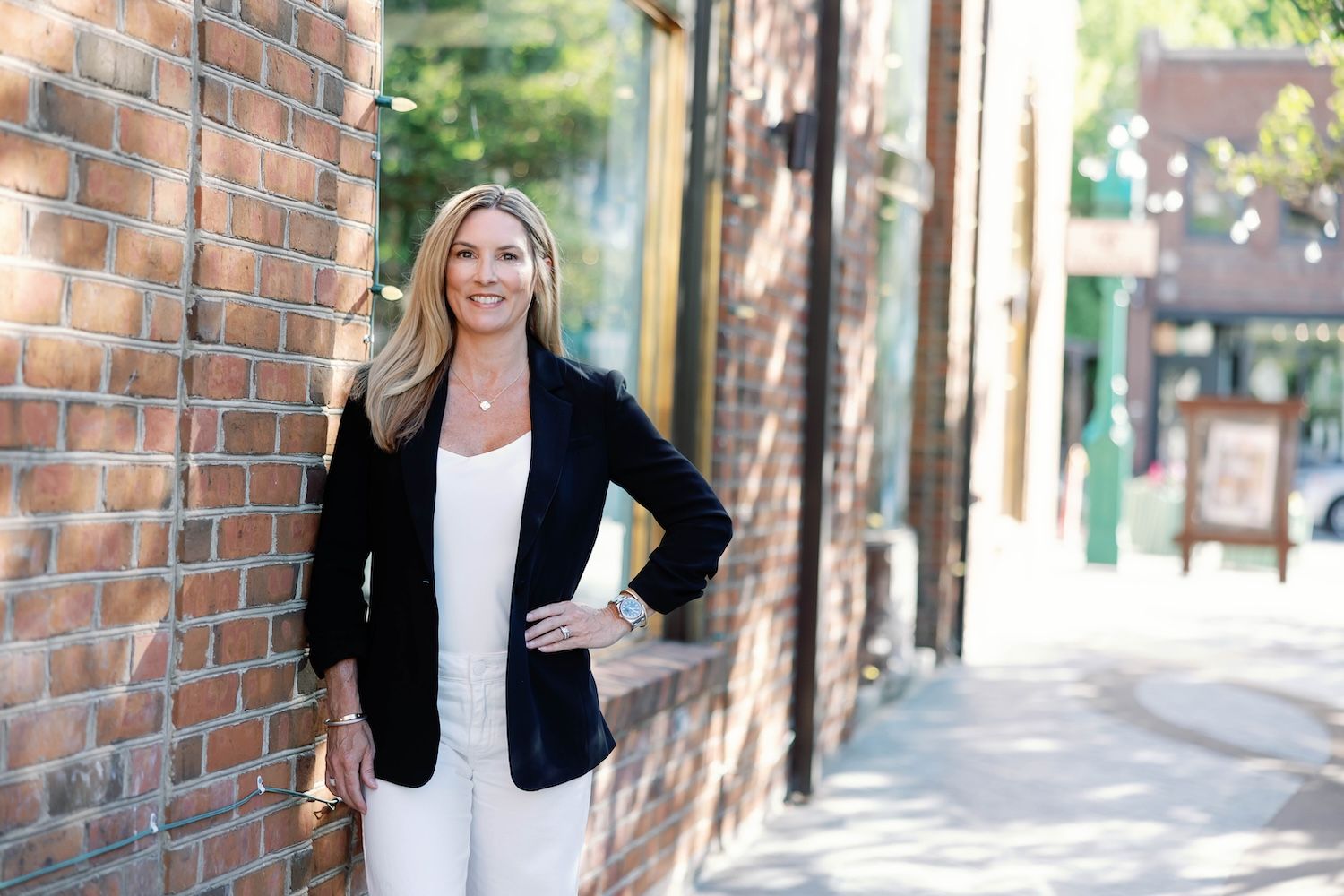 Jennifer LeMaster in smart casual attire smiling by a brick wall on a sunny street.