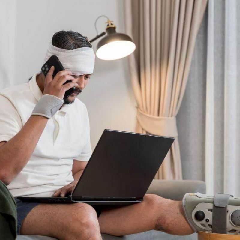 Man with a bandaged head and a leg in a cast sits on a sofa, talking on a phone while using a laptop nearby at home.