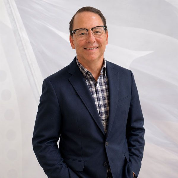 Smiling man in a navy blazer and plaid shirt, standing against a light abstract background.