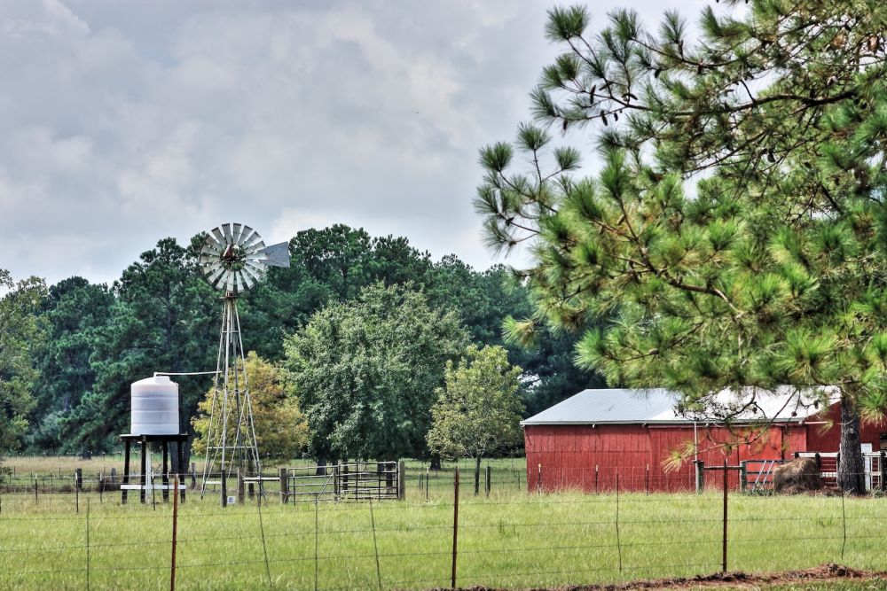 Rural farm scene with windmill, red barn, and green pasture under a cloudy sky.