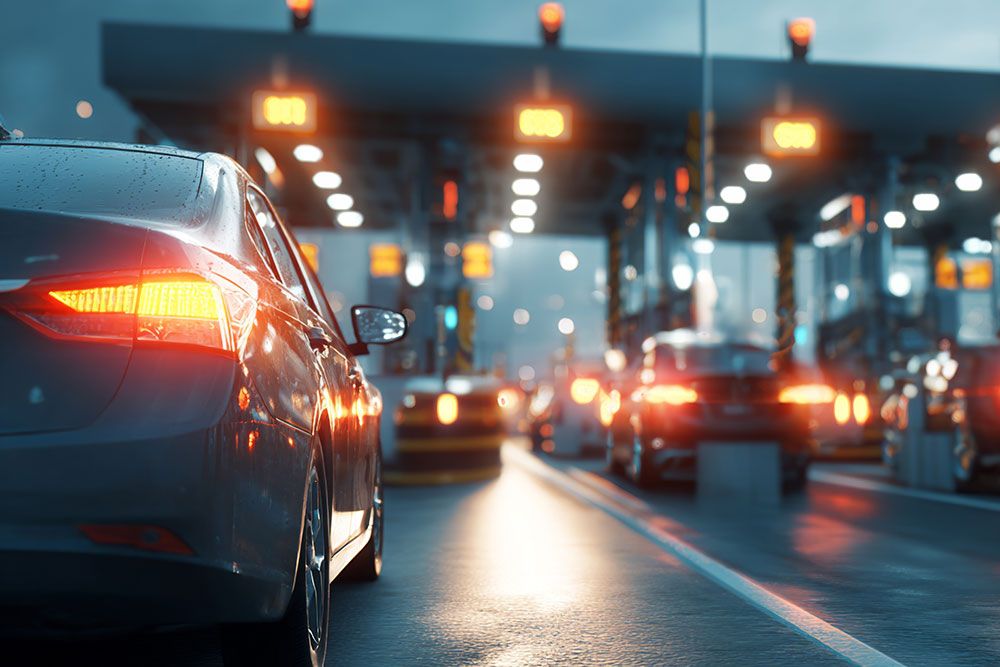 Cars queue at illuminated toll booths during evening, with brake lights casting reflections on wet road.