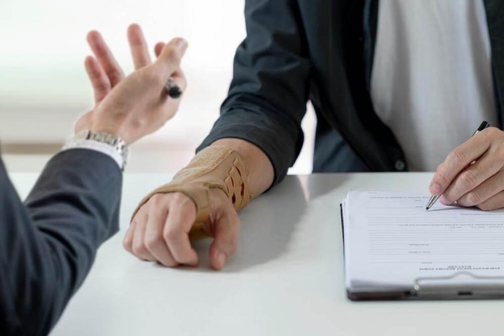 Handshake across a table between two people, one with a forearm in a beige cast, a pen and forms on a clipboard nearby.