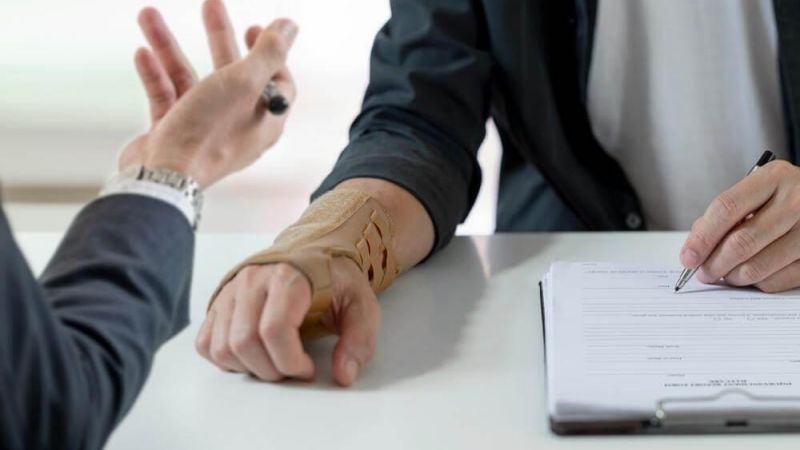 Handshake across a table between two people, one with a forearm in a beige cast, a pen and forms on a clipboard nearby.