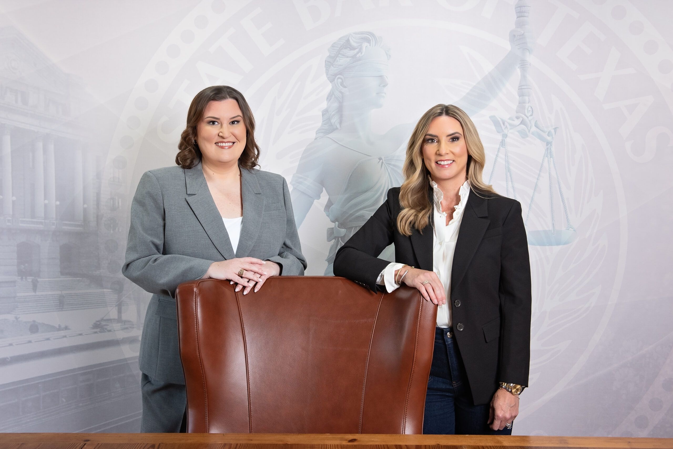 Two women in professional attire standing behind a conference chair at a legal office with a Texas seal backdrop.