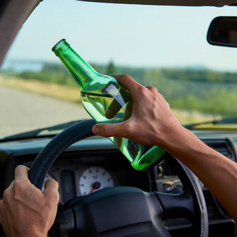Person holding green bottle while driving, emphasizing dangers of drinking and driving on the road.