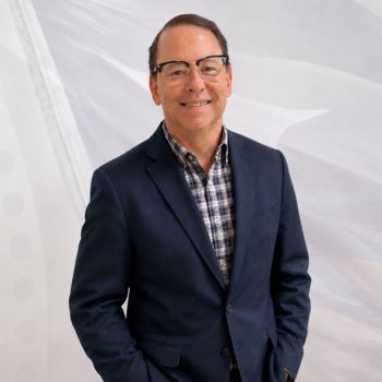 Smiling man in a navy blazer and plaid shirt, standing against a light abstract background.