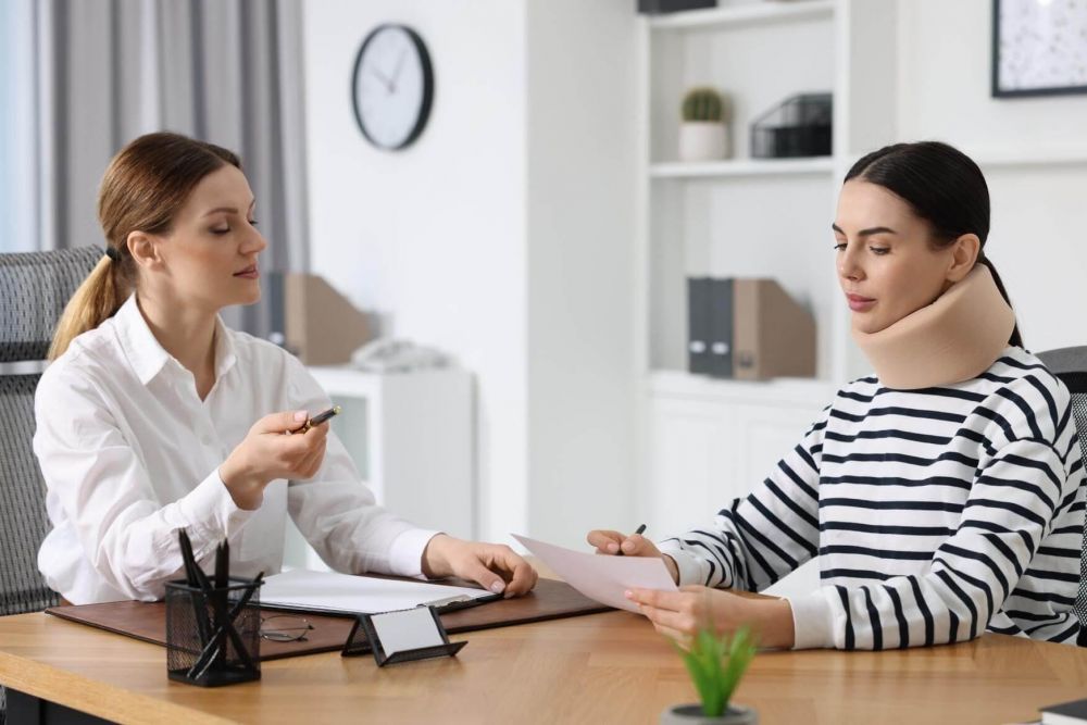 Clinician explains documents to a patient wearing a neck brace in a bright office.